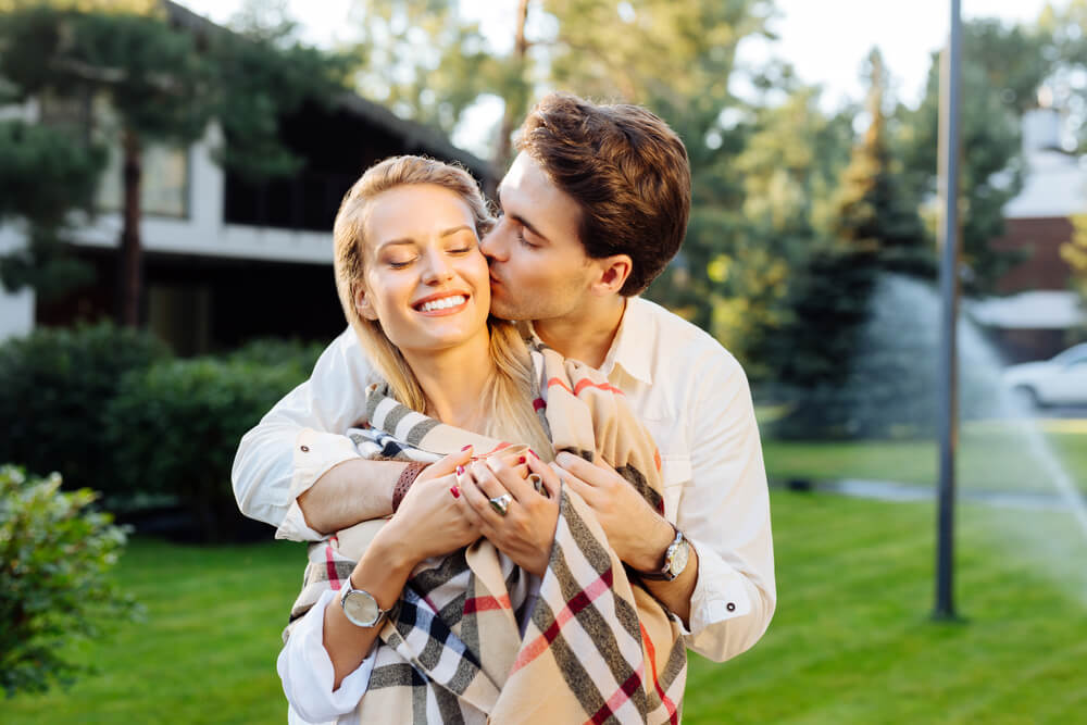 A couple. The man is embracing the woman from behind and kissing her. Tips on dating a widower: 6 things you need to know. Click here.