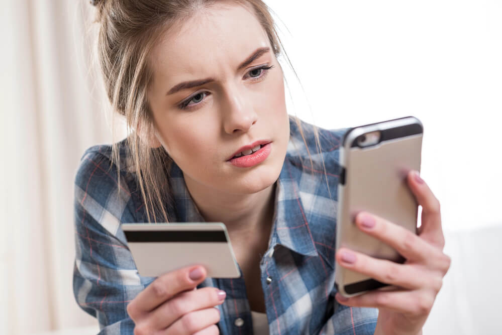 A woman checking her mobile phone while holding a card with the other hand. Here are tips on how to get free gas and discount gas in 2024: 8 legit ways.
