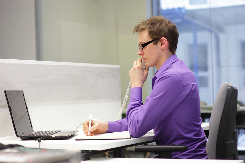 Office worker sitting upright at a desk while using a laptop, telephone, and notebook