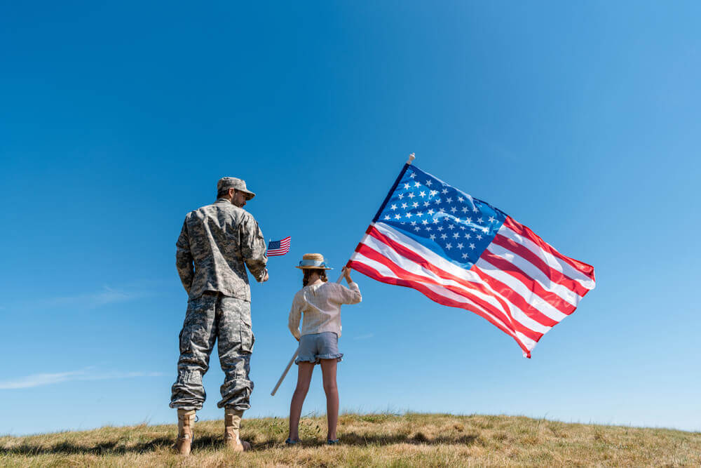 Soldier in military uniform standing near kid with american flag against blue sky