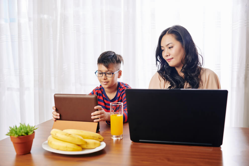 A person working on a laptop at a kitchen table with a child nearby
