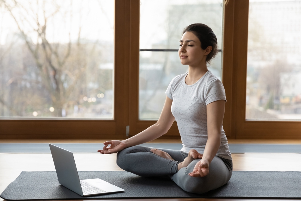 Woman sitting cross-legged on a yoga mat beside an open laptop
