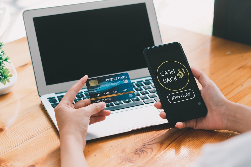 Hands holding a phone displaying cash back beside a laptop
