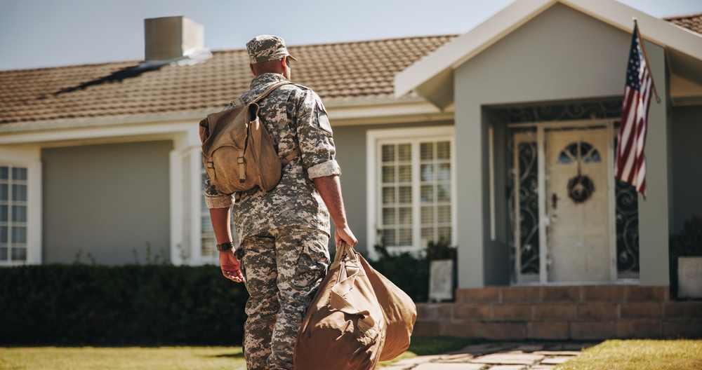 Serviceman in camouflage carrying luggage toward a house with an American flag