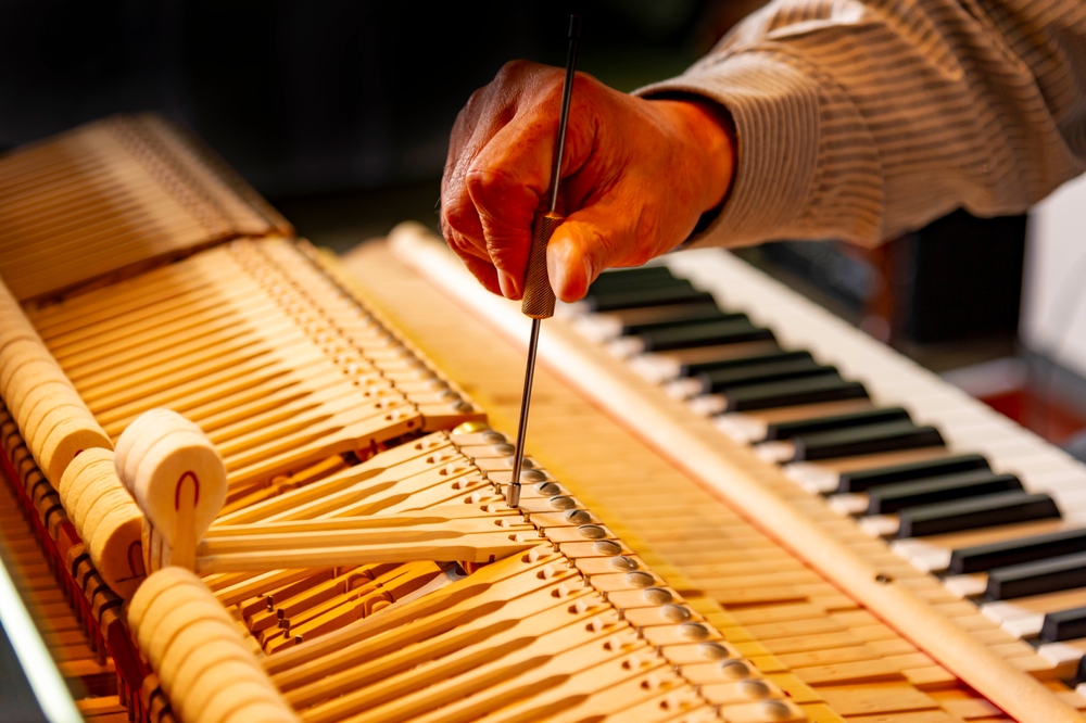 Piano technician using a precision tool to adjust the exposed internal mechanism beside the keys