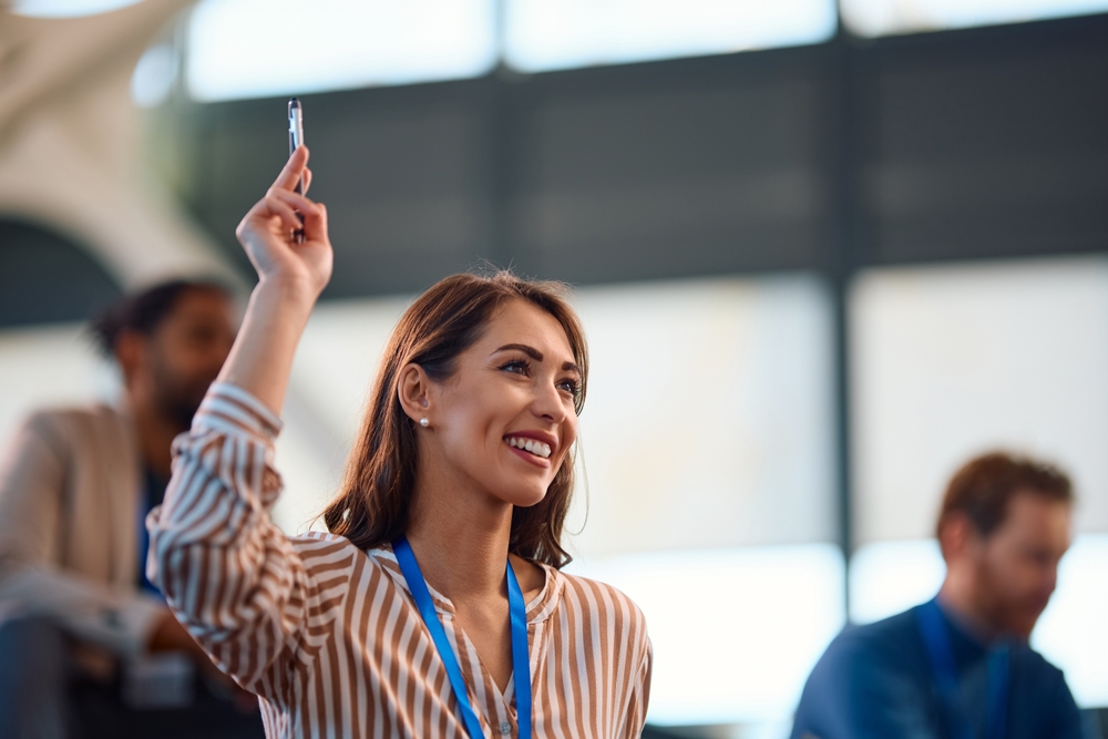 Woman wearing a conference lanyard raising her hand to ask a question among seated attendees