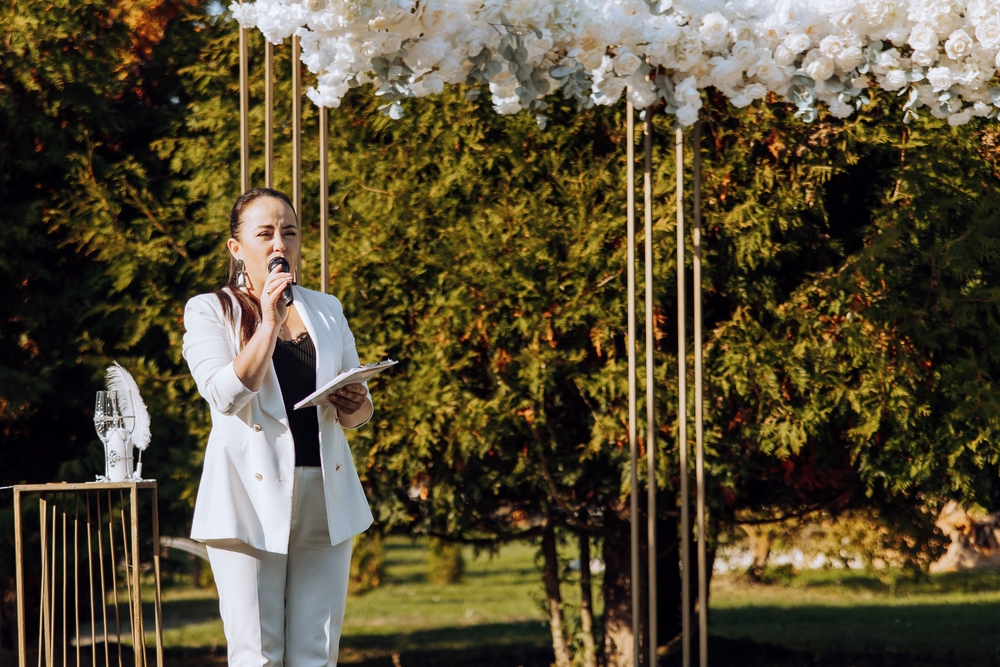Woman in a white suit reading from a folder and speaking into a microphone beside an outdoor wedding arch