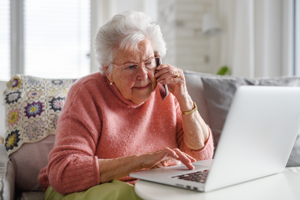 Older woman talking on a phone while using a laptop at home