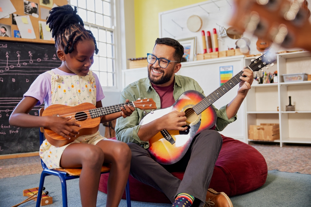 Music teacher playing an acoustic guitar beside a child practicing a ukulele during a lesson