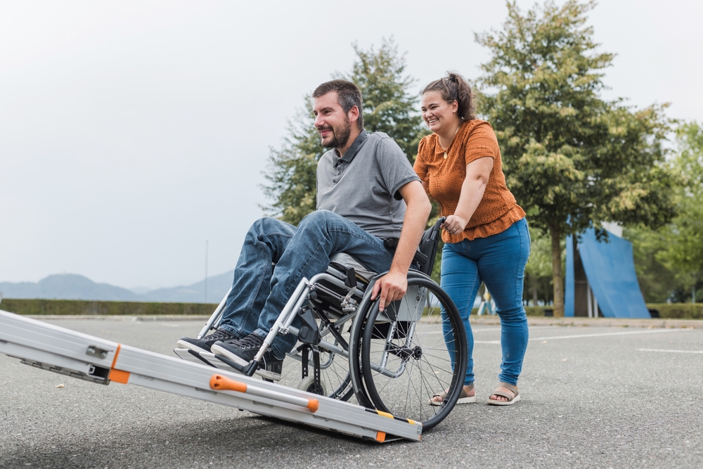 Woman assisting a wheelchair user up a portable vehicle ramp outdoors