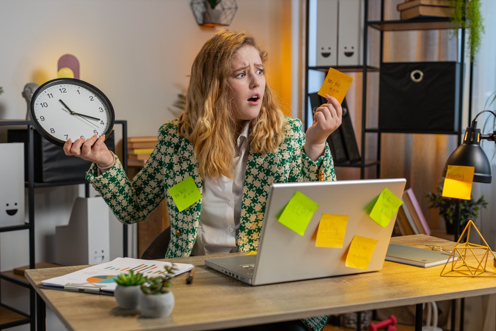Overloaded worker holding a clock and sticky note beside a laptop covered in tasks