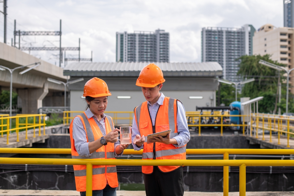 Two workers wearing hard hats examining a water sample beside treatment tanks