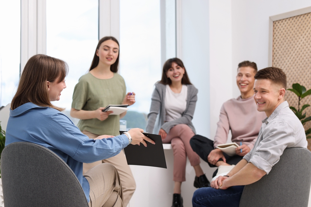 Woman facilitating a seated group discussion while participants listen with notebooks