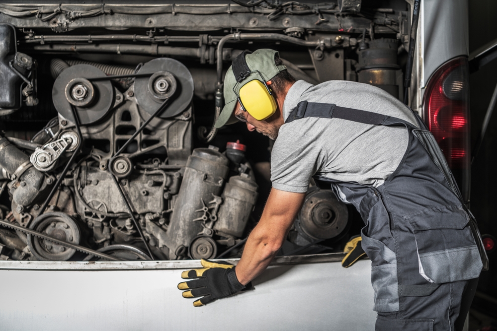 Mechanic wearing hearing protection and gloves working in the open rear engine bay of a bus
