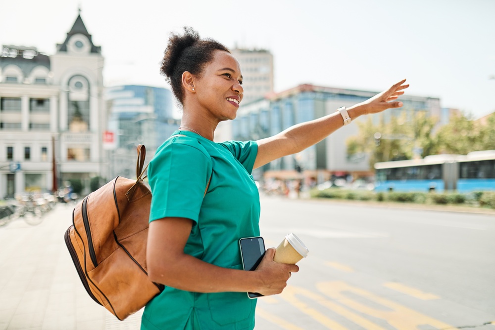 Smiling woman in green medical scrubs carrying a backpack and coffee outside