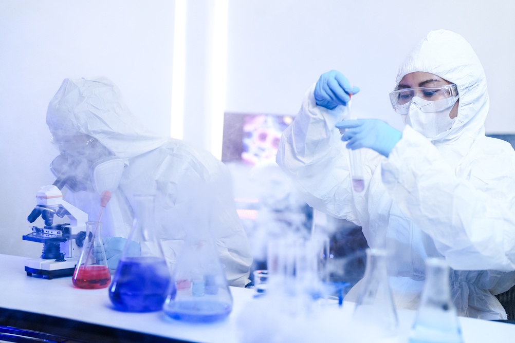 Laboratory researchers wearing protective suits and goggles examine fluid samples beside a microscope