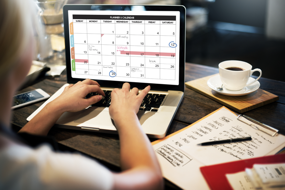 Person managing a laptop calendar beside a clipboard checklist and coffee