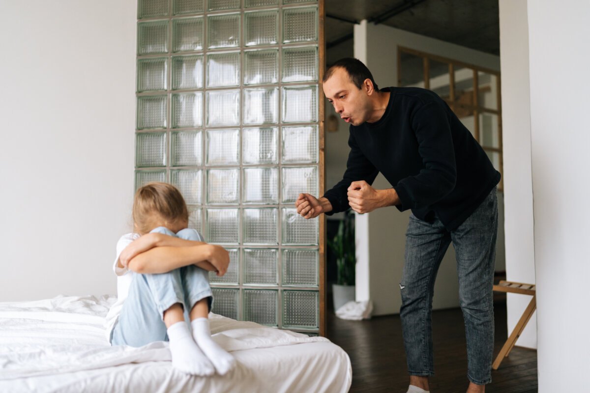 Standing aggressive young father scolding, raising voice, shouting at depressed stubborn difficult little child daughter sitting on bed hugging knee.