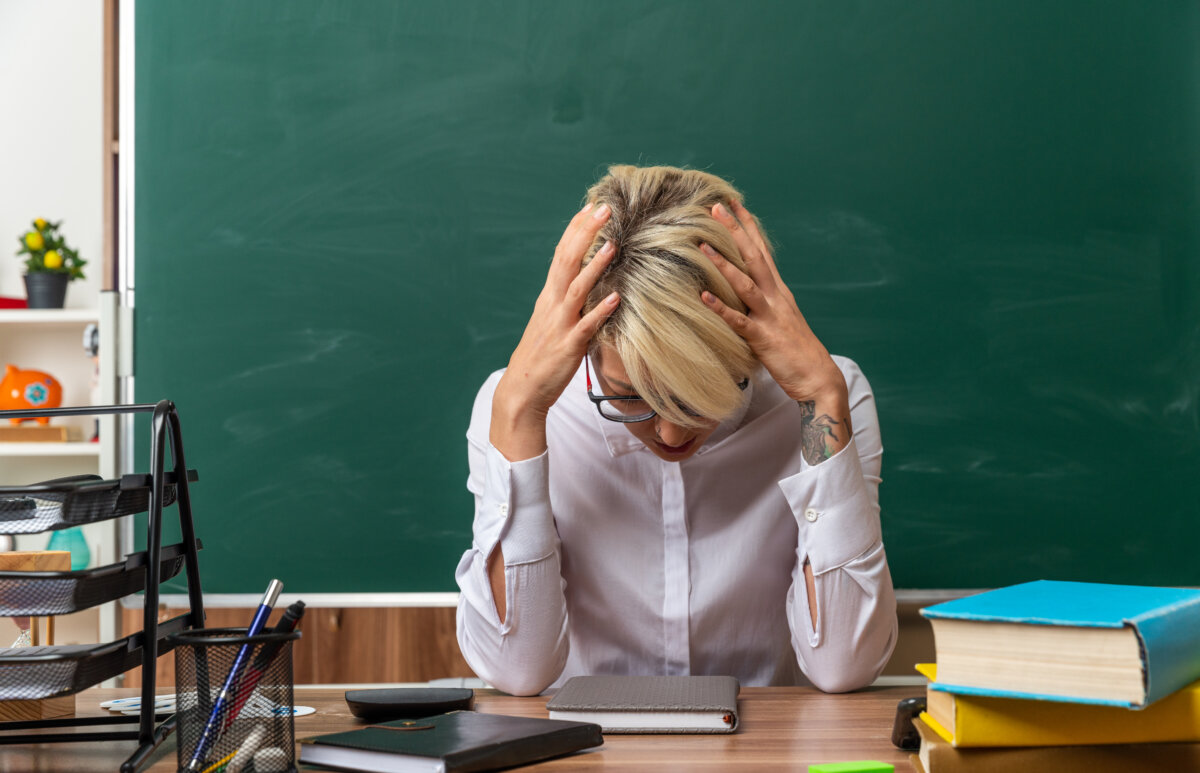 stressed young blonde female teacher wearing glasses sitting at desk with school tools in classroom keeping hands on head